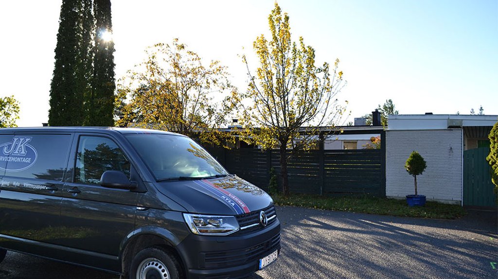 En mörkgrå servicebil från JK Service & Montage parkerad framför ett bostadshus på en solig dag. Bilen används för installation och service av kyl- och värmepumpar, vilket framgår av logotypen på sidan.